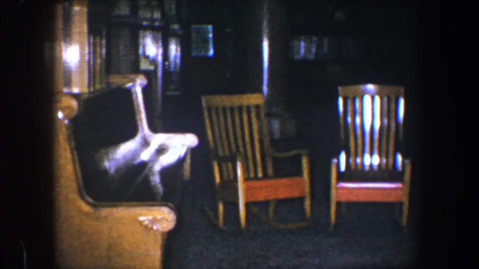 1960 Baltimore 8mm frame with a closer view of an armchair, wooden rocking chairs, dark wall surfaces, and carpet in low light.