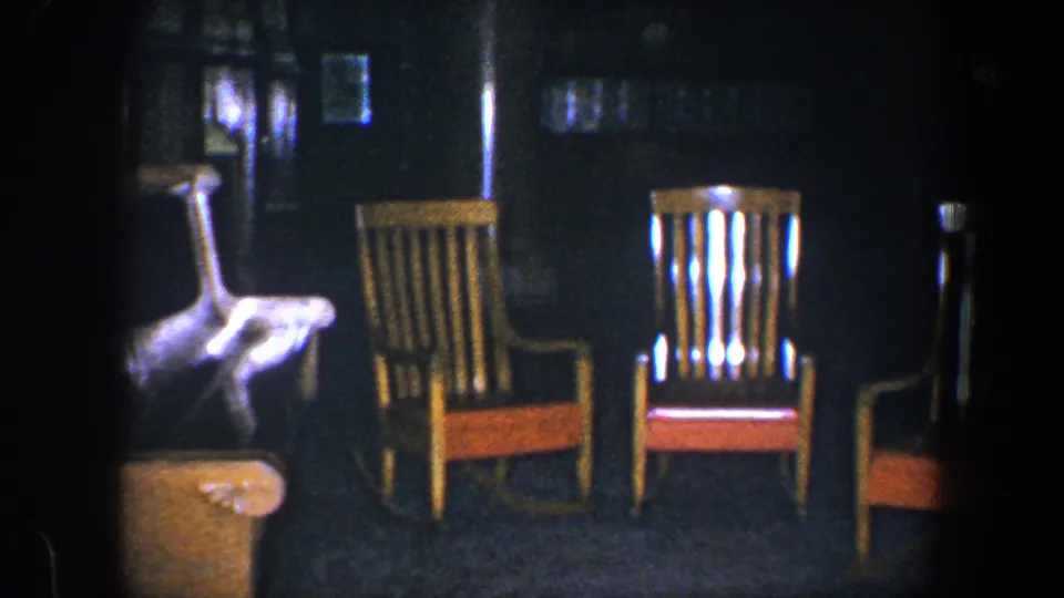1960 Baltimore 8mm frame showing two wooden rocking chairs, the edge of an upholstered armchair, and a shadowed interior wall.