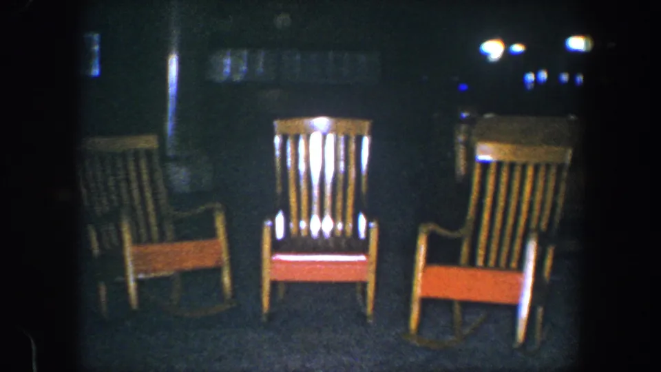 1960 Baltimore 8mm frame with three wooden rocking chairs in low light, backed by faint reflections and dark room details.
