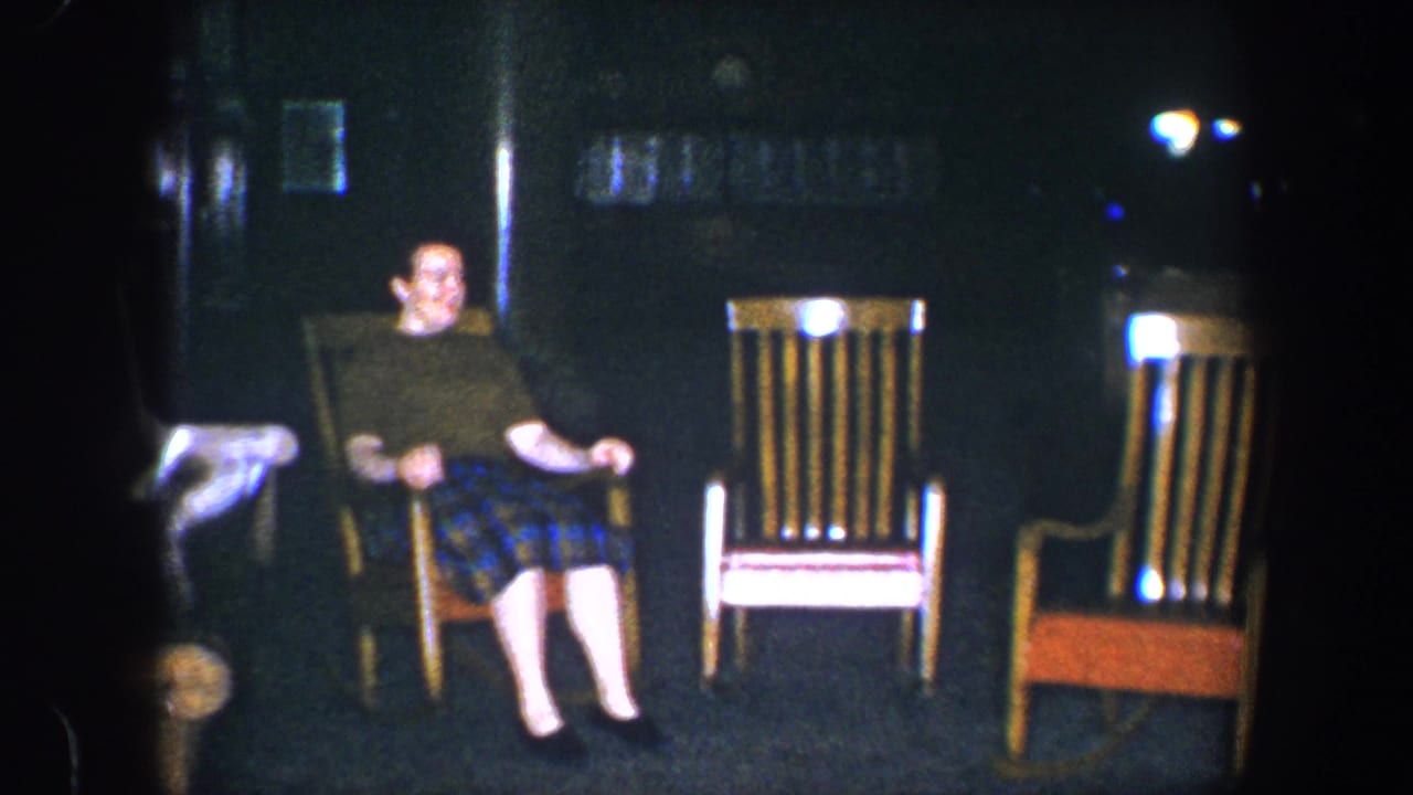 Adult woman in a green shirt and plaid skirt sits in a wooden rocking chair in a dim Baltimore home interior, 1960 metadata supplied.