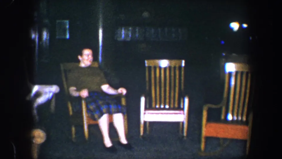 Adult woman in a green shirt and plaid skirt sits in a wooden rocking chair in a dim Baltimore home interior, 1960 metadata supplied.