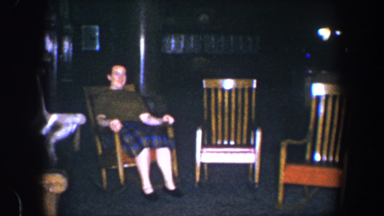 The woman remains seated in the rocking chair, with two empty wooden chairs visible in the dim 1960 Baltimore interior.