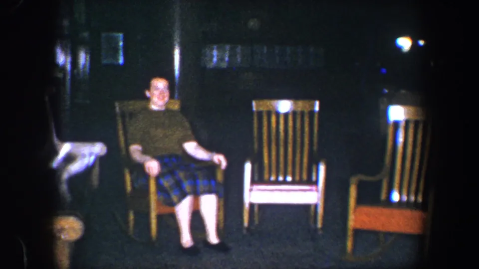 A fixed 8mm view shows the woman in a green shirt and plaid skirt seated among wooden rocking chairs in a dark room.
