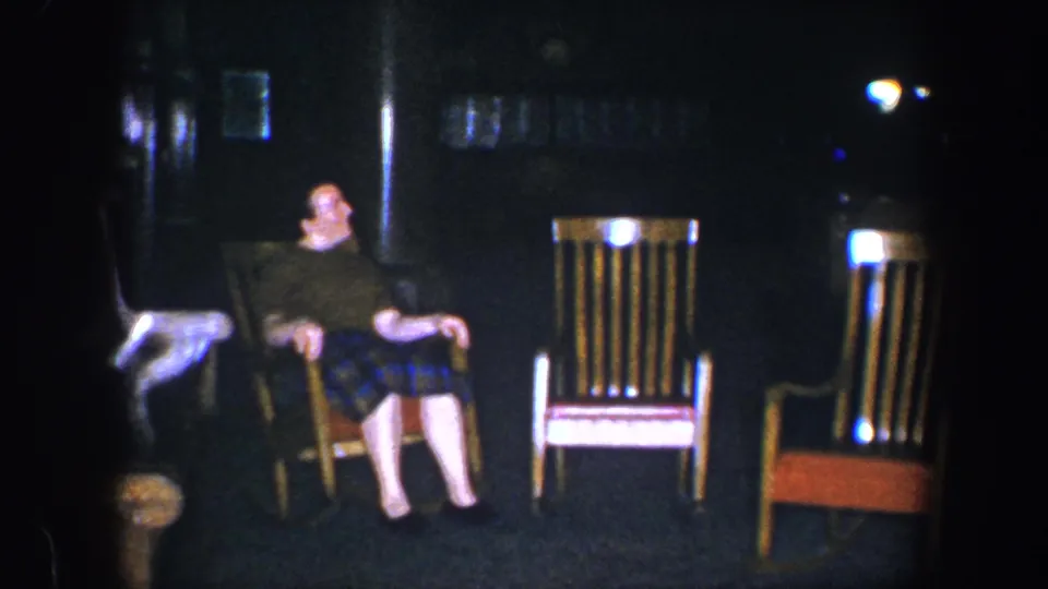 The 8mm frame shows the man leaning back in his chair in the dim 1960 Baltimore interior, with empty wooden chairs beside him.