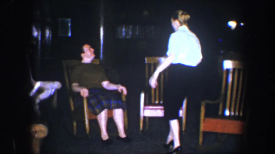 A woman in a light shirt and dark skirt passes in front of the seated man, adding candid movement to the 1960 Baltimore interior scene.