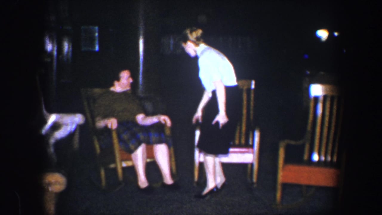 Two women near wooden chairs in a dim room, one seated and one leaning forward while standing.