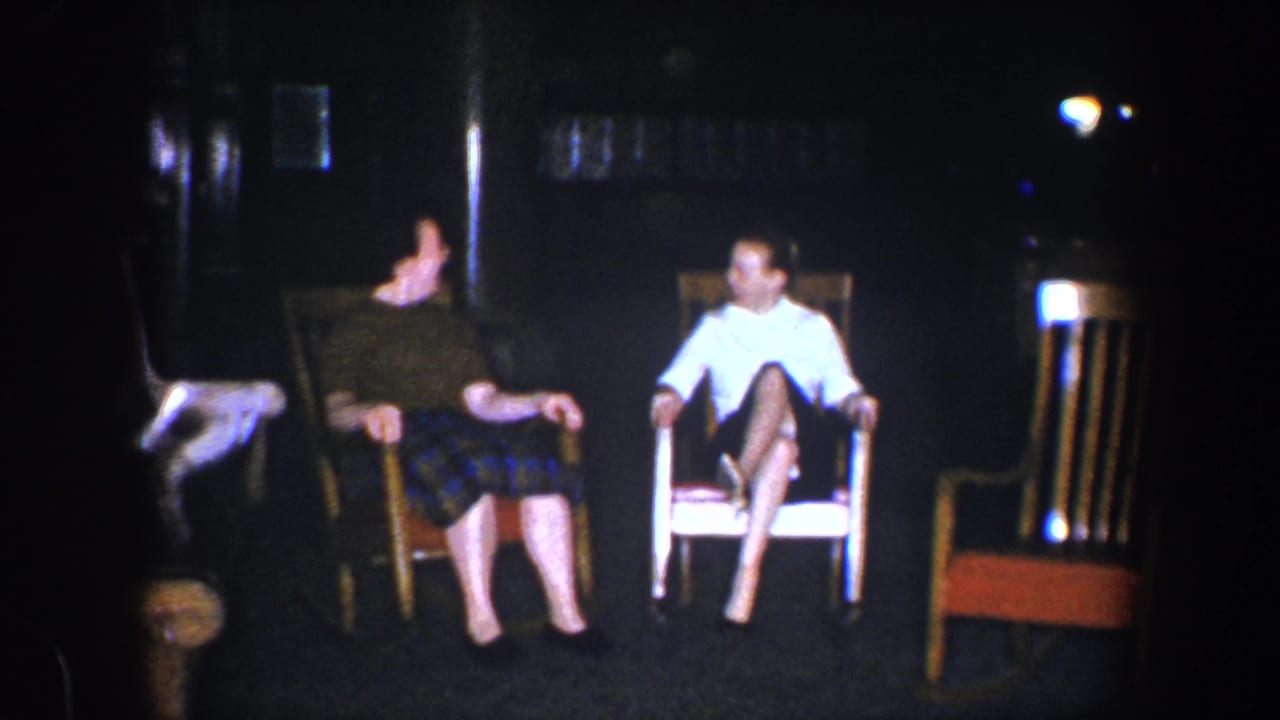 Two adult women sit side by side in a low-lit Baltimore home interior, with wooden chairs and dark wall surfaces behind them.
