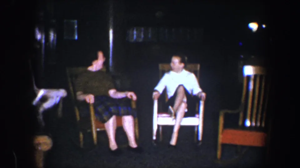 Two adult women sit side by side in a low-lit Baltimore home interior, with wooden chairs and dark wall surfaces behind them.