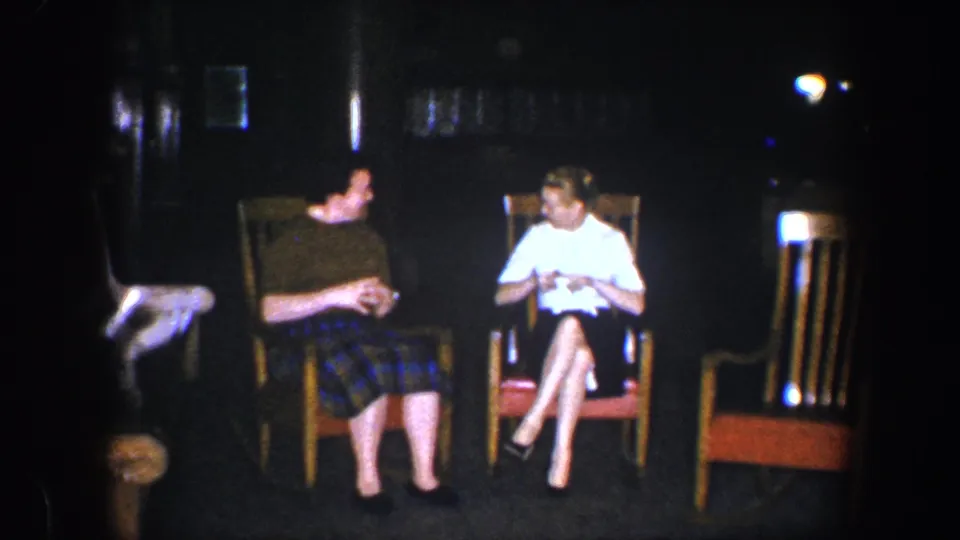 Two women appear mid-conversation in a 1960 Baltimore home-movie interior, seated in wooden chairs with soft focus and dark surroundings.