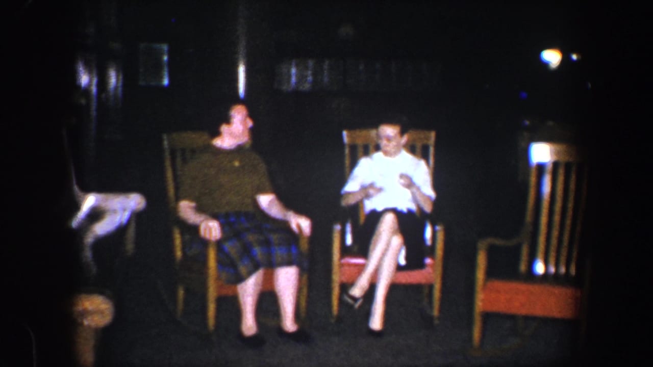 Two women sit in wooden rocking chairs in a dim interior, with an empty chair nearby.