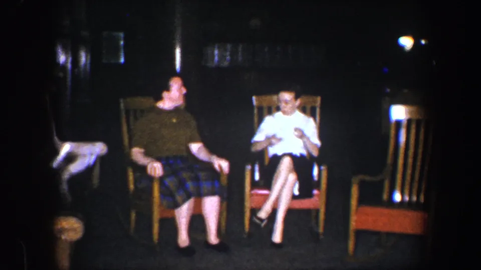 In 1960 Baltimore home-movie footage, two adult women sit in wooden rocking chairs in a dark room, one in a plaid skirt and the other in a white blouse and dark skirt.