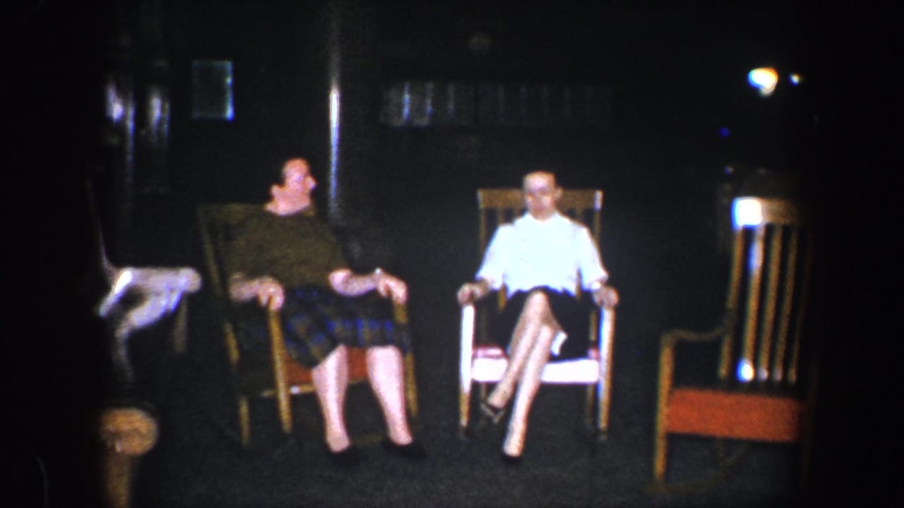Two seated women face forward in rocking chairs as one rests her arms on the chair.