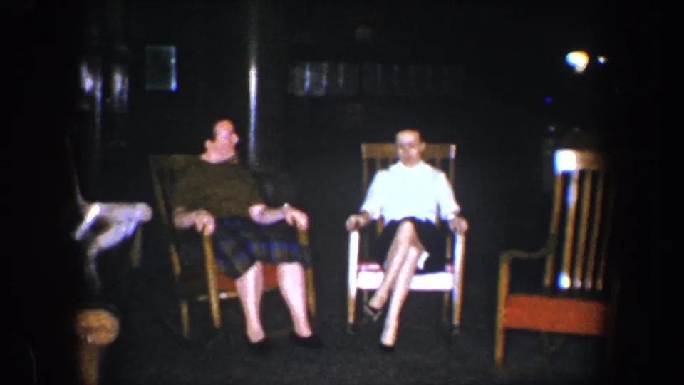 The two women remain seated in wooden rocking chairs, with the dark-shirted woman in a plaid skirt and the woman in white sitting with crossed legs in the dim interior.