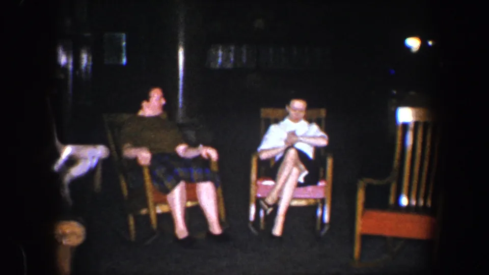 During the 1960 Baltimore home-movie clip, the woman in the dark top turns toward the woman in the white blouse as they sit close together in wooden rocking chairs.