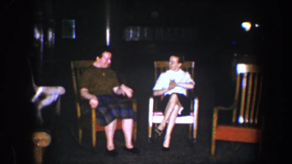 The women appear to converse in the dim private interior, seated in rocking chairs with an additional empty wooden chair visible at the right edge of the frame.