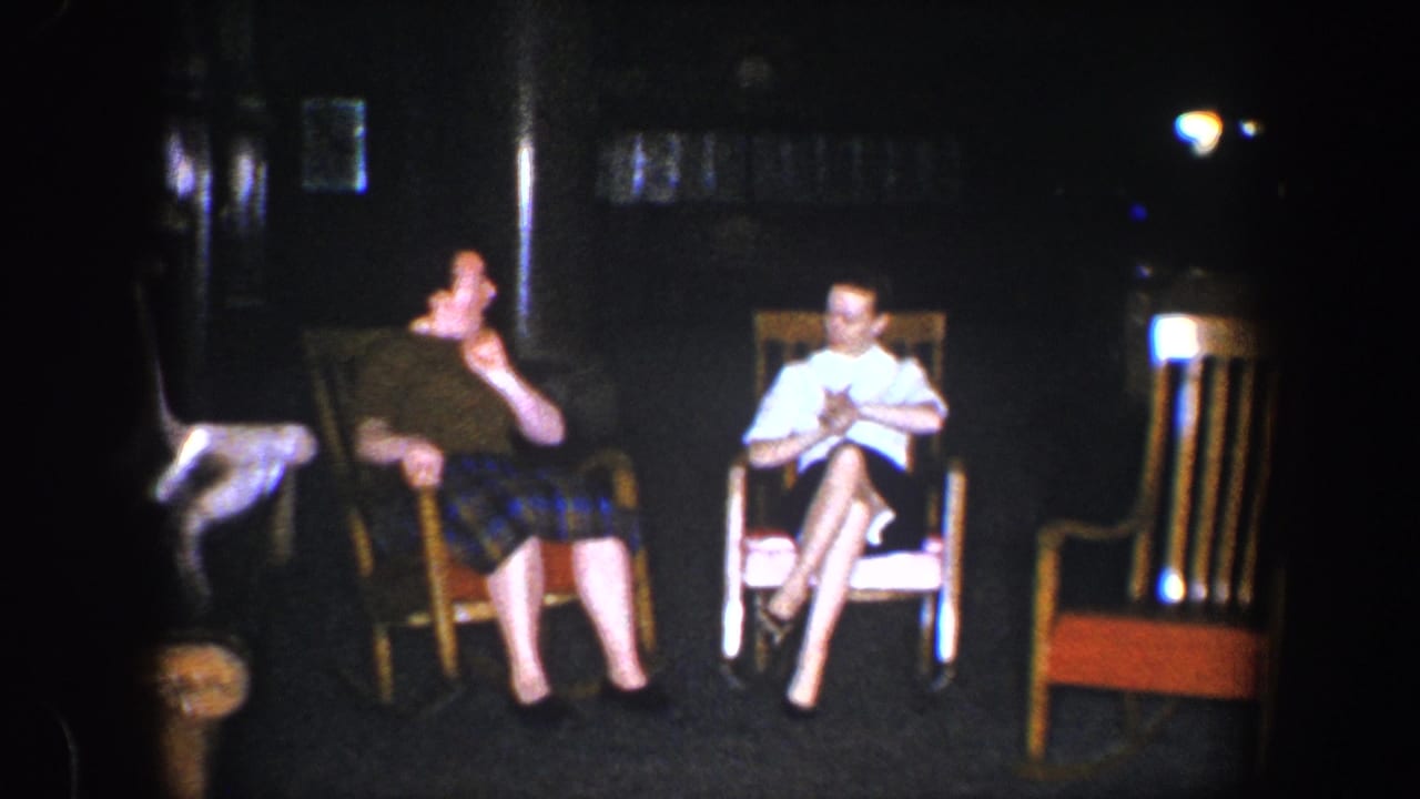 1960 Baltimore 8mm frame showing two young women seated in wooden rocking chairs in a dark indoor room.