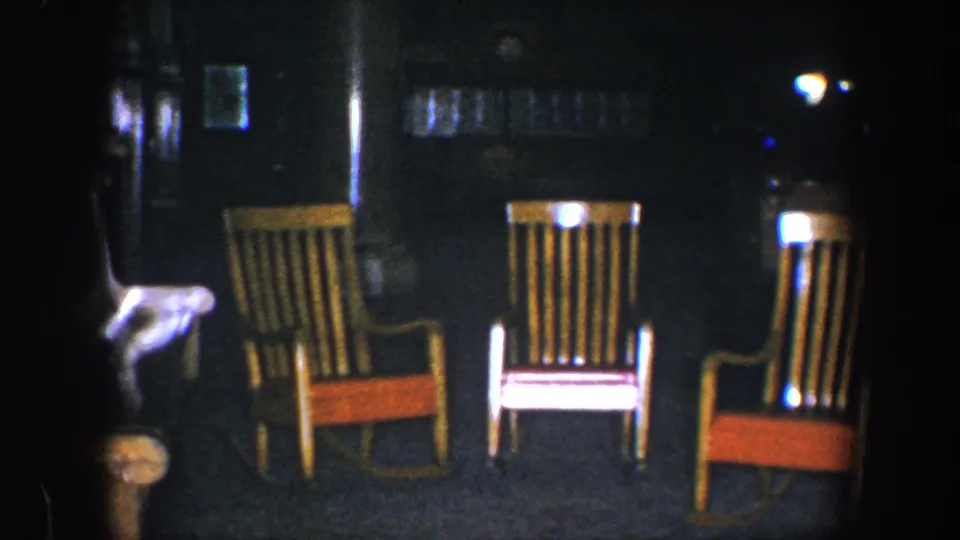 1960 8mm Baltimore frame with three wooden rocking chairs in soft focus, orange seat panels, and a dark surrounding interior.