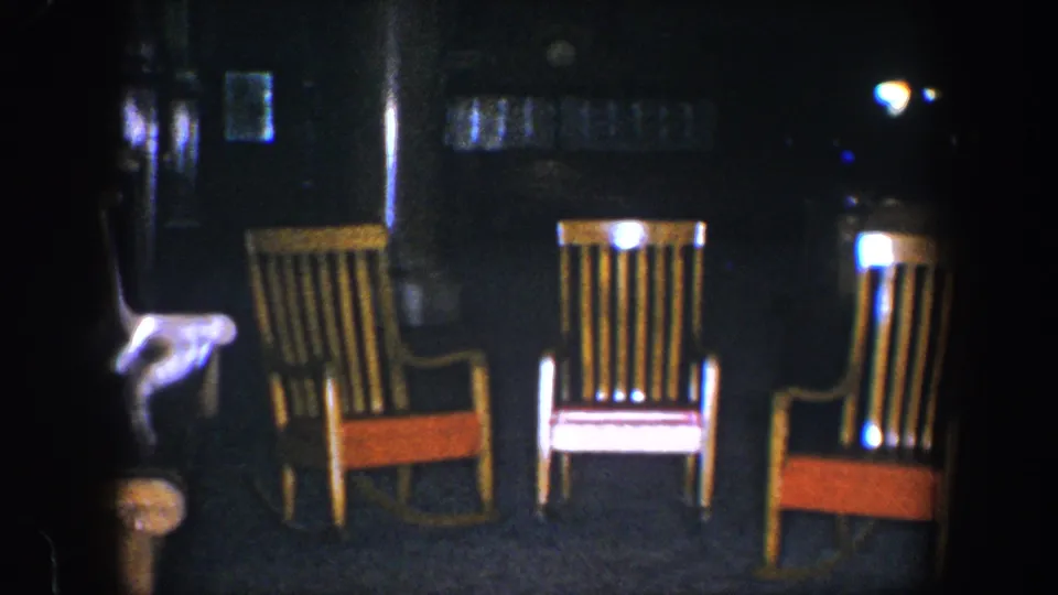 Baltimore 1960 home-movie frame showing three orange-seated rocking chairs in a dim domestic interior with visible film grain.