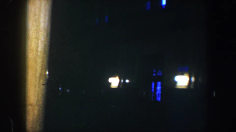 The 1960 Baltimore home-movie frame holds on a dark exterior area with a vertical post and faint blue-white lights beyond it.