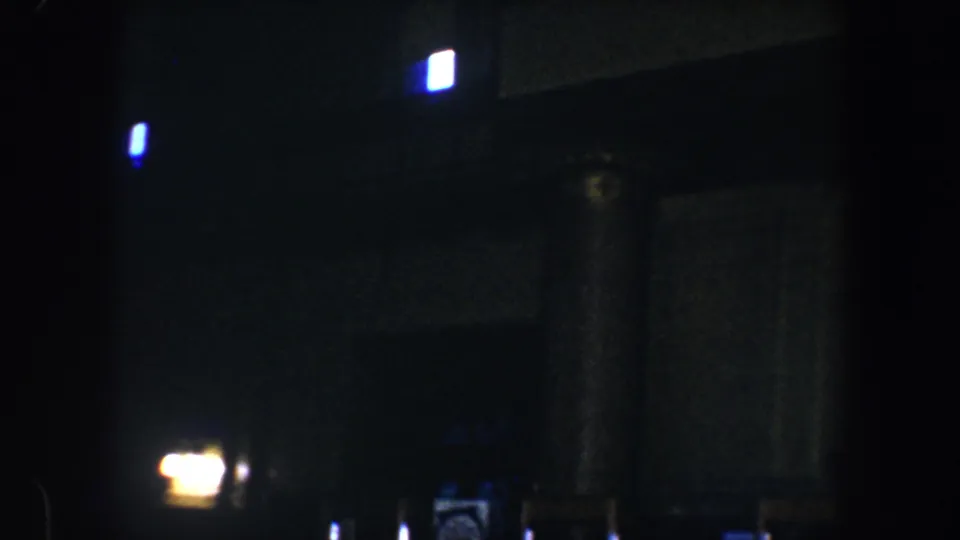 Low-light 1960 Baltimore 8mm frame showing a dark interior with glowing lights, columns, a doorway, signs, and a wall clock.