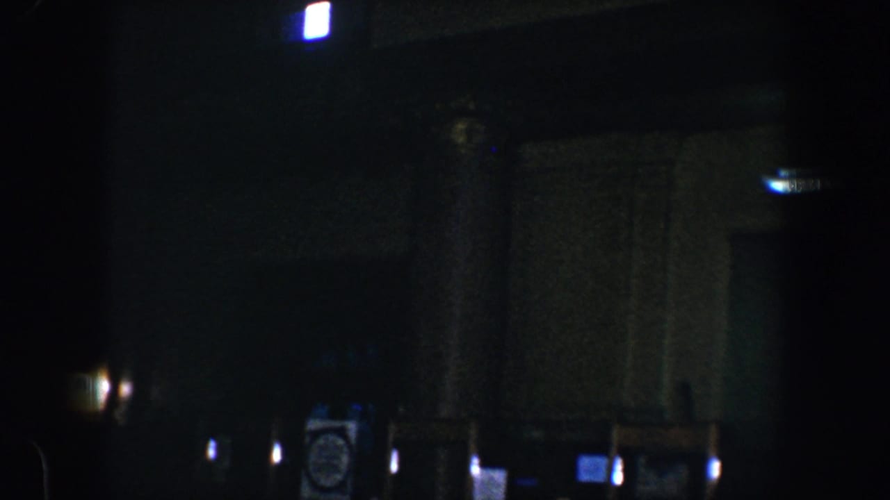 Shadowed interior with bright doorway light, columns, clock, signs, and dark wall surfaces.
