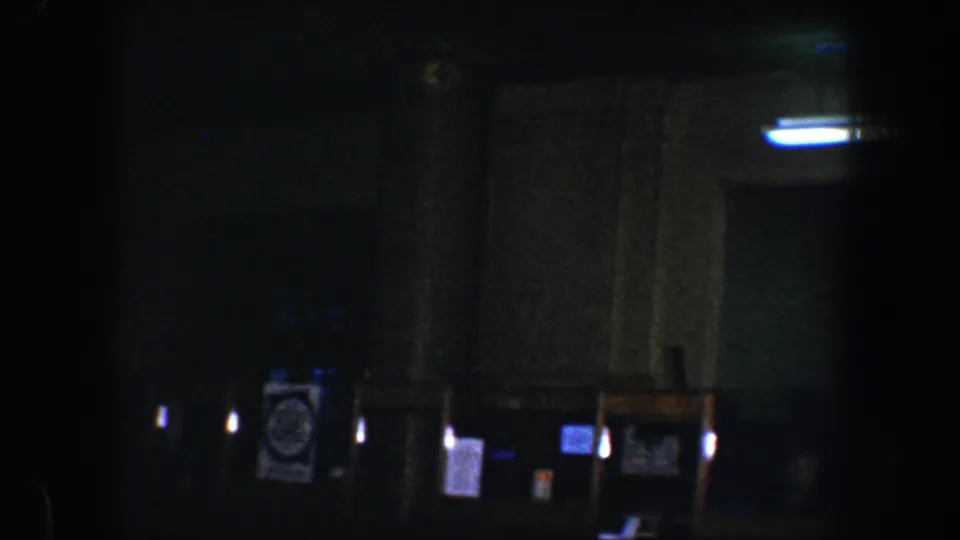 Low-exposure 1960 Baltimore 8mm frame with dark columns, a doorway, signs, small glowing lights, and possible figures in shadow.
