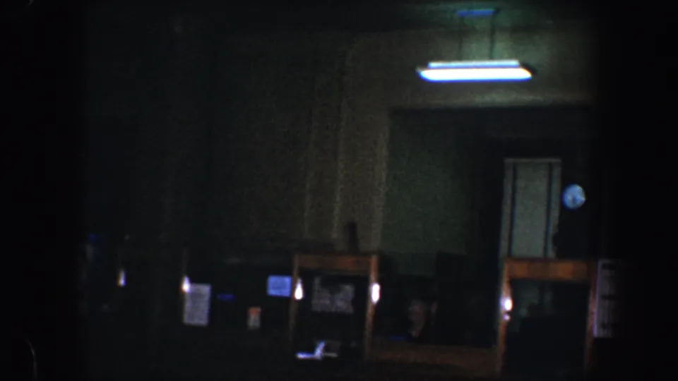 1960s 8mm home-movie frame of a dim Baltimore interior with a fluorescent light, doorway, clock, and shadowed counter area.