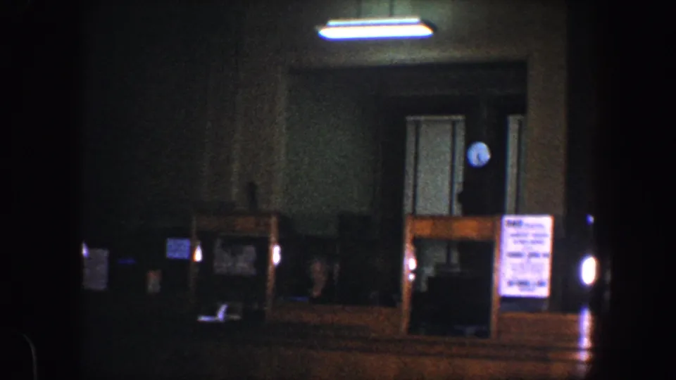 A dark 1960s 8mm frame of a Baltimore room with fluorescent lighting, a doorway, counter surface, posted notice, and clock.