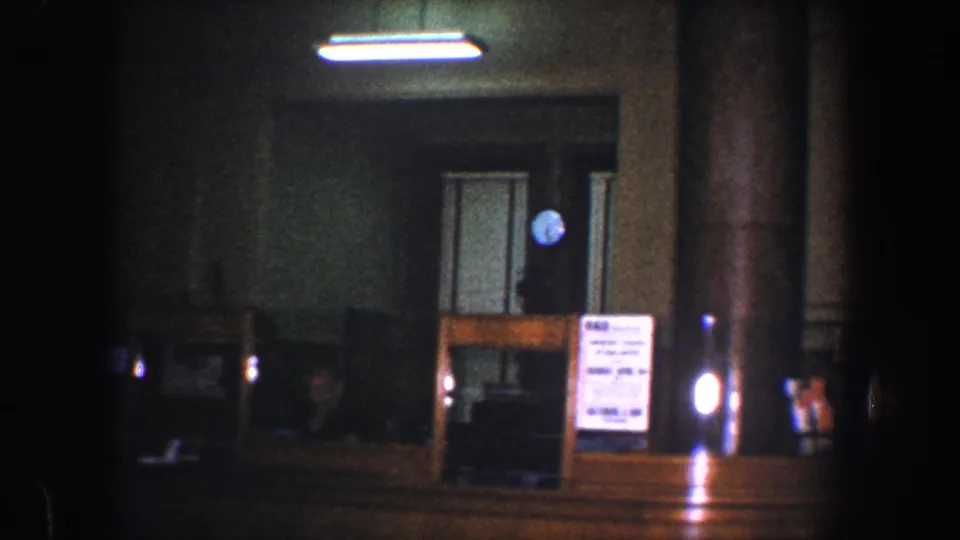 1960s Baltimore home-movie frame with a dim interior, fluorescent ceiling light, doorway, counter, posted notice, and a small person at the right edge.