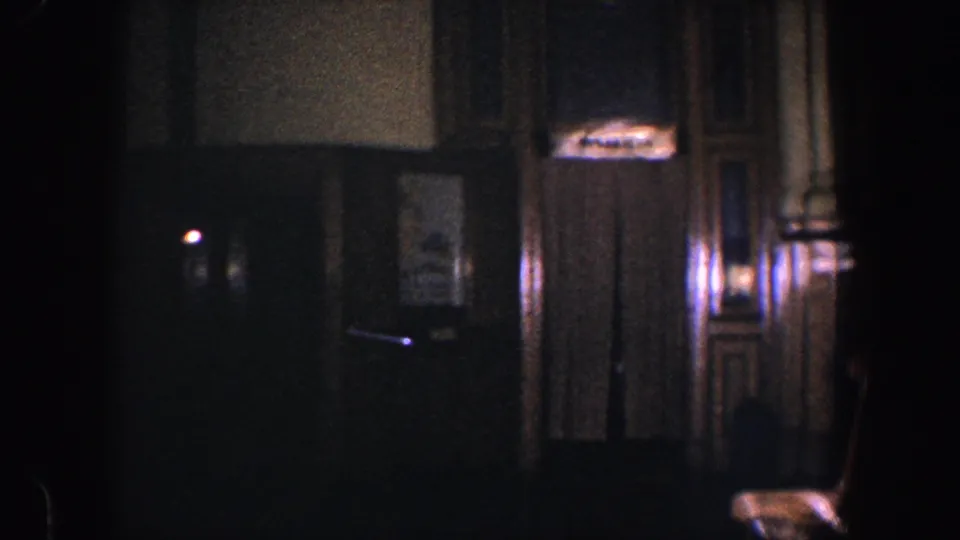 1960 Baltimore home-movie frame showing a dark interior hallway with wood paneling, curtained elevator area, sign, and chair.