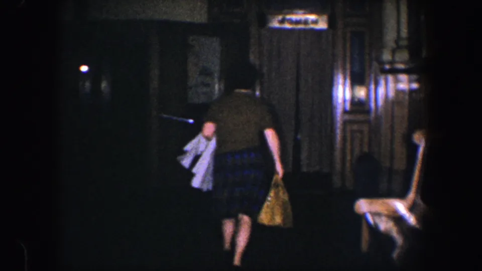 1960 Baltimore home-movie frame of a woman walking toward an elevator area with a yellow bag and white garment in hand.