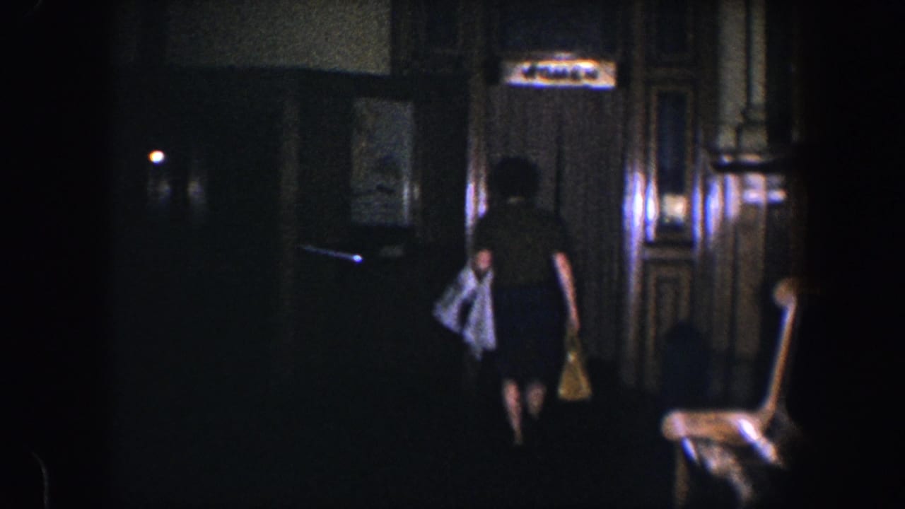 Woman seen from behind walking through a dim interior with a coat and handbag.