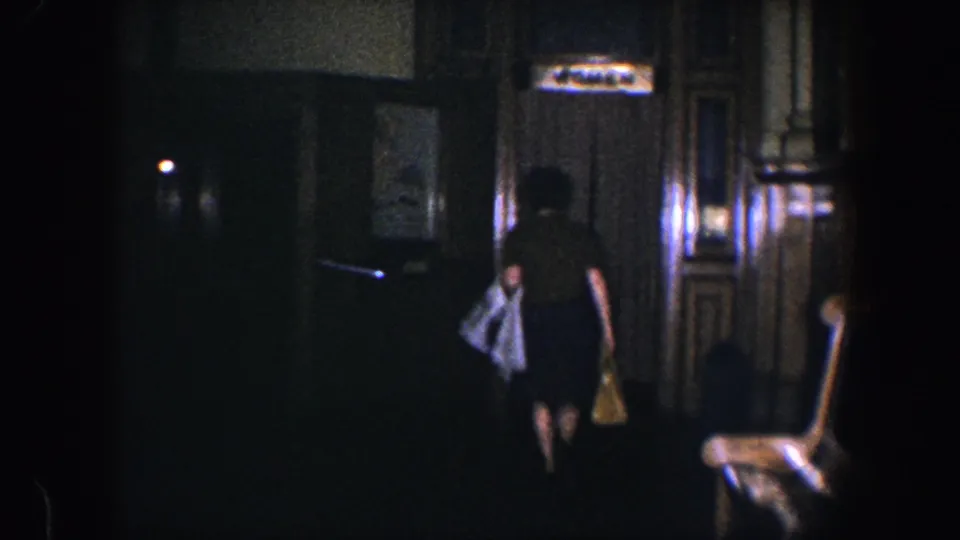 In a 1960 Baltimore home interior, a woman walks away from the camera past dark wood paneling while carrying a coat and handbag.