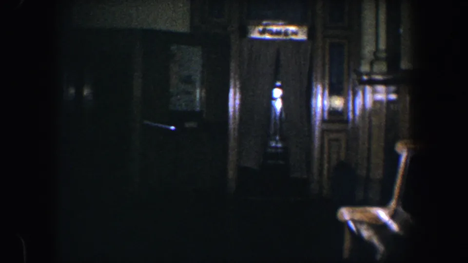 The dim Baltimore home interior is nearly empty of visible figures, showing curtains, paneled trim, wall art, and a chair in low 8mm exposure.
