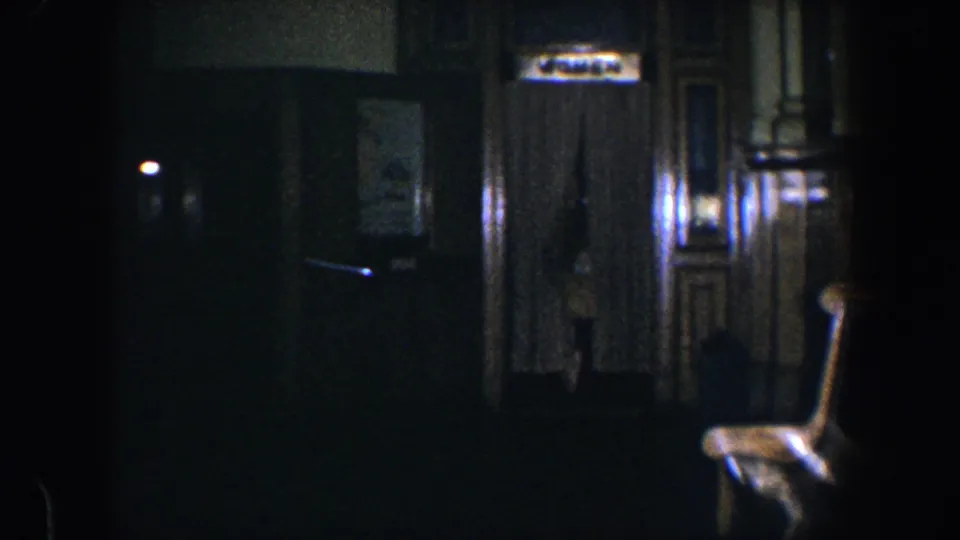 A woman is faintly visible near the curtained doorway of a dim 1960 Baltimore private home interior, with wood paneling and a chair nearby.