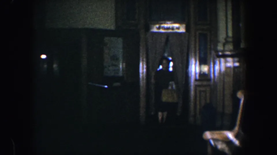 A woman stands or moves near a doorway in a dim 1960 Baltimore home interior with dark paneling and wall lighting.