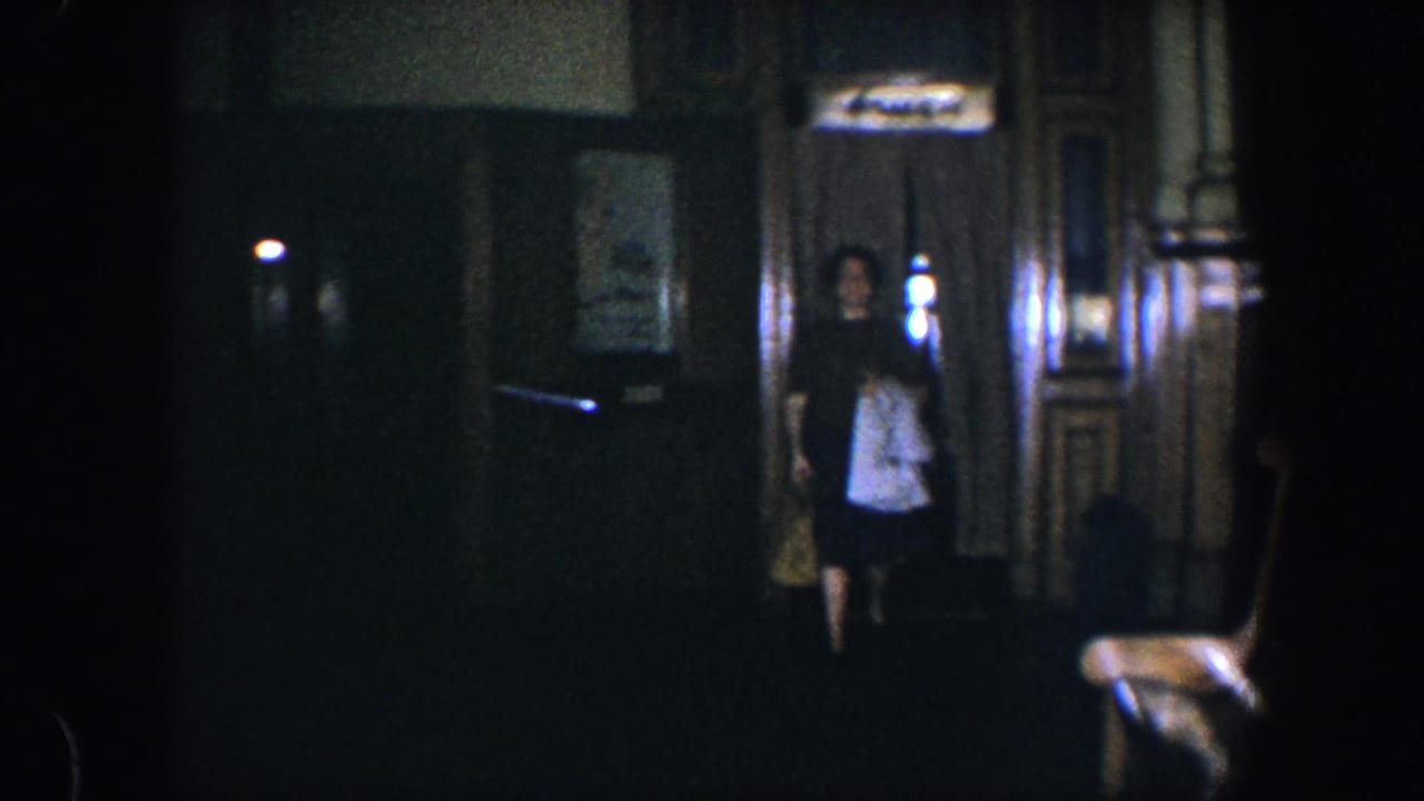 Woman walking into a dark paneled room while holding a light garment and bag.