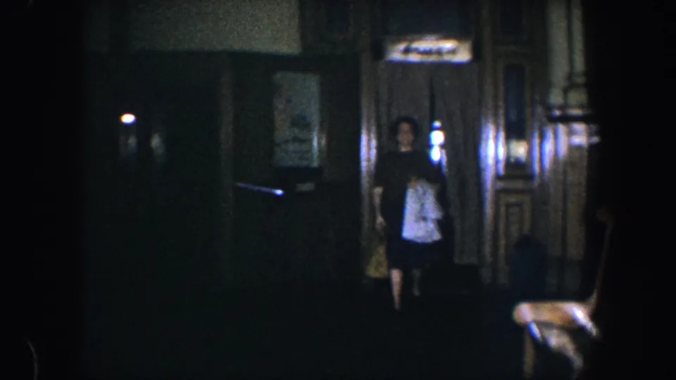 The woman enters a dim Baltimore home interior, carrying a light-colored garment and handbag against dark wood paneling.