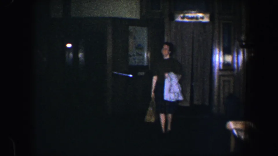 A woman walks toward the camera in the dim 1960 Baltimore interior, holding a handbag and light draped garment.