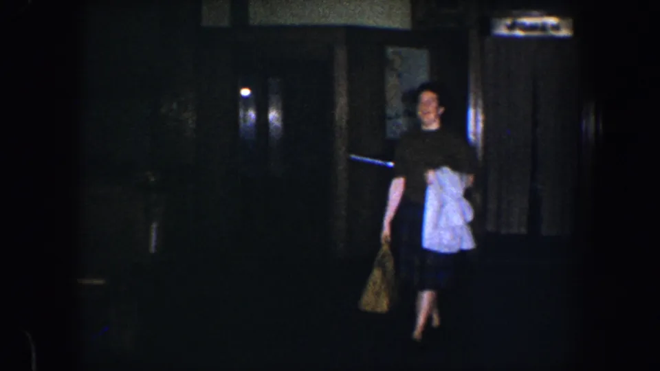 The woman continues across the dim Baltimore home interior, framed by dark woodwork and soft wall lighting.