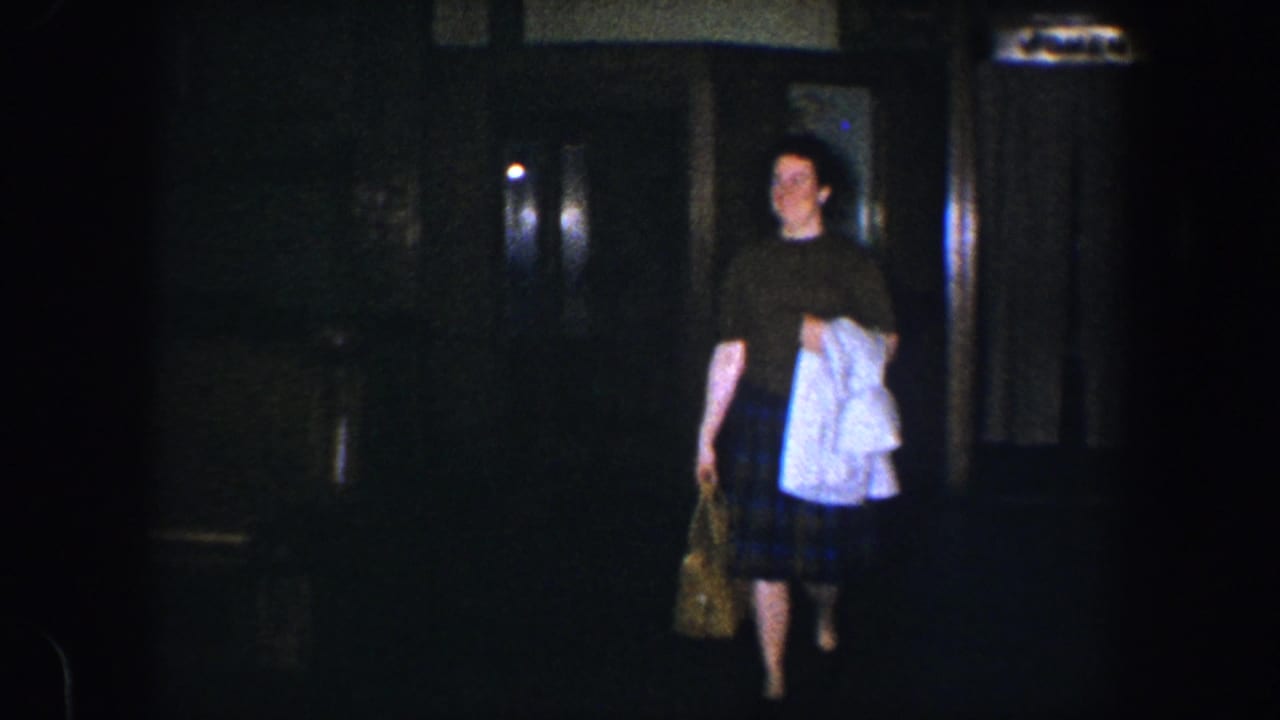 Woman walking through a dark interior while carrying a white garment and patterned bag.