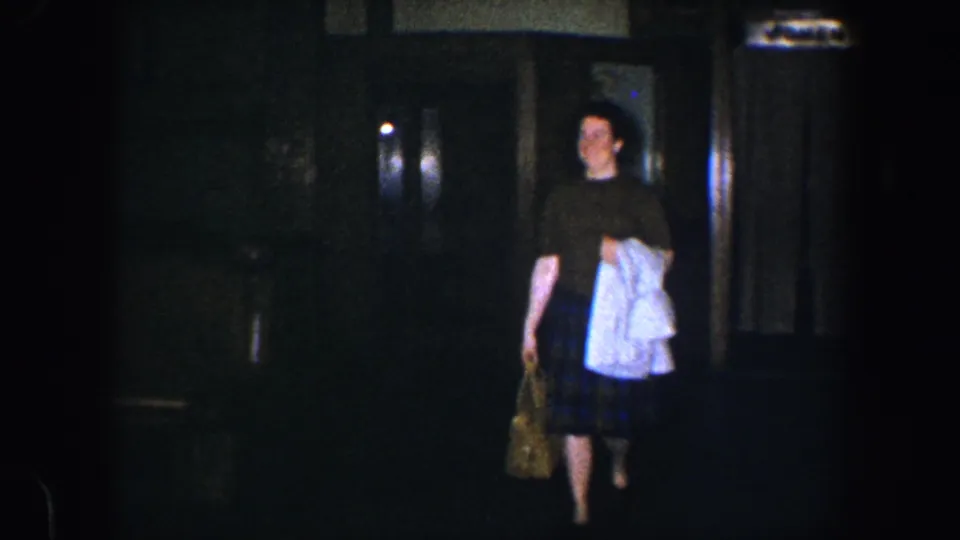 In 1960 Baltimore home-movie footage, a woman in a brown top and plaid skirt walks through a dim interior with clothing and a bag.