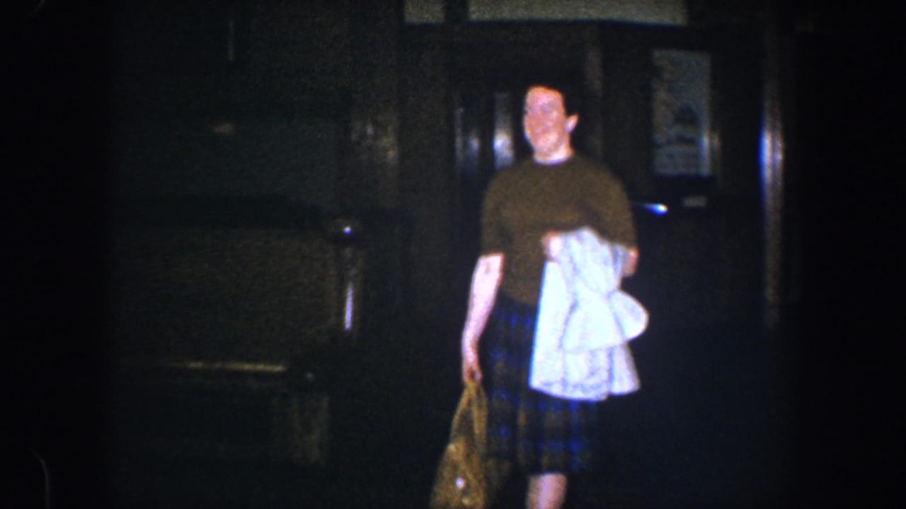 The woman smiles as she nears the camera in the dim interior, holding a white garment and patterned bag in 1960 Baltimore footage.