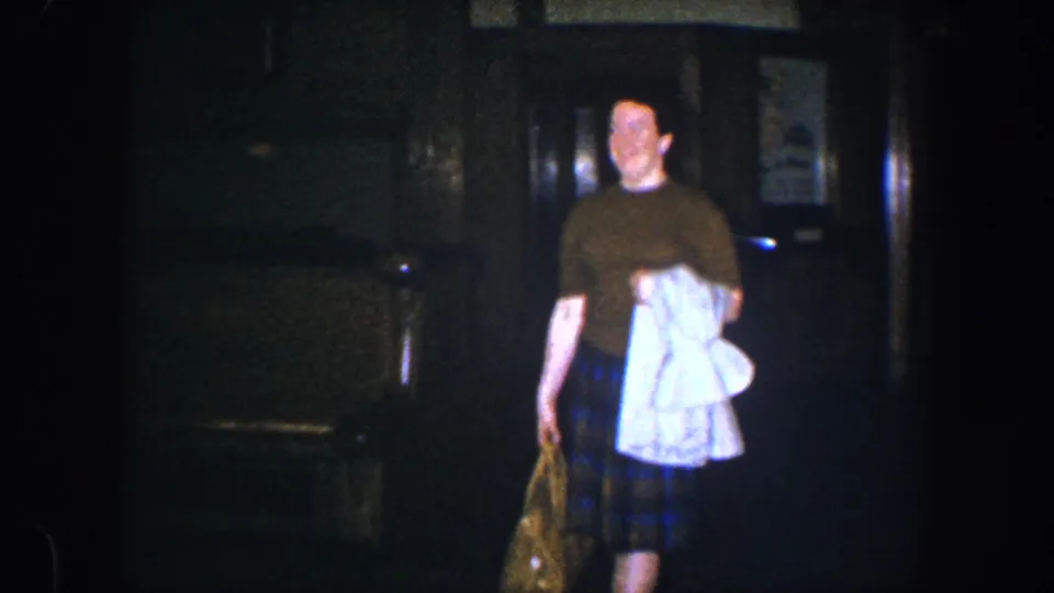 The woman smiles as she nears the camera in the dim interior, holding a white garment and patterned bag in 1960 Baltimore footage.