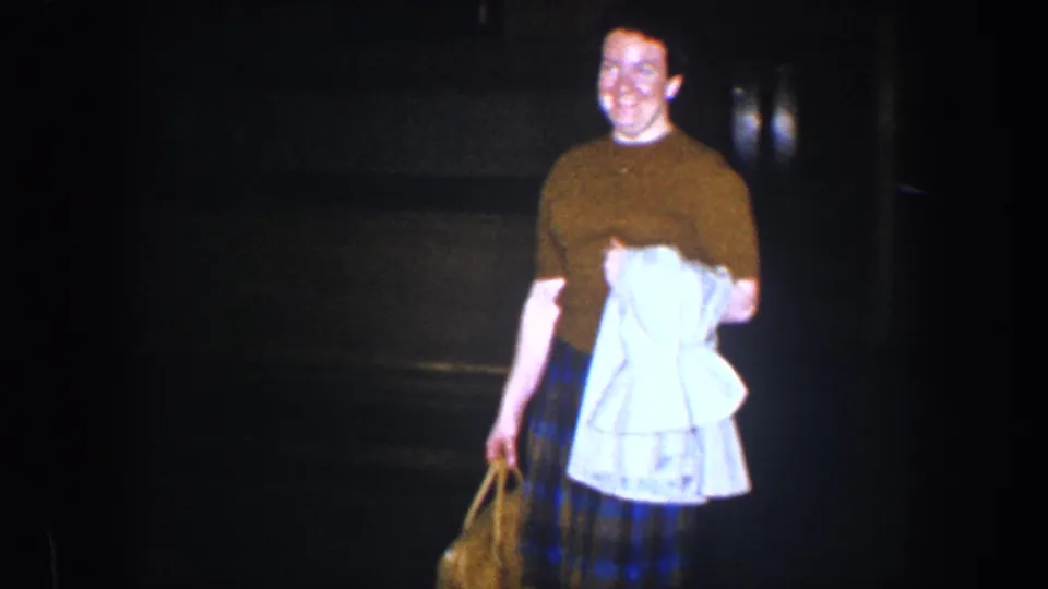 A close home-movie view shows the woman smiling in a brown top and plaid skirt while carrying clothing and a bag indoors.