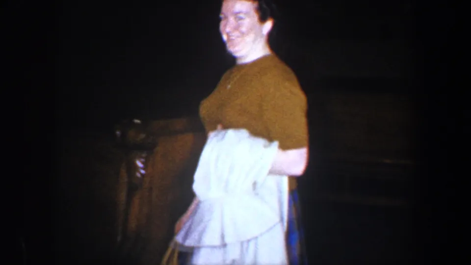 Near the end of the shot, the smiling woman passes close to the camera inside a dark Baltimore interior, holding white clothing and a bag.
