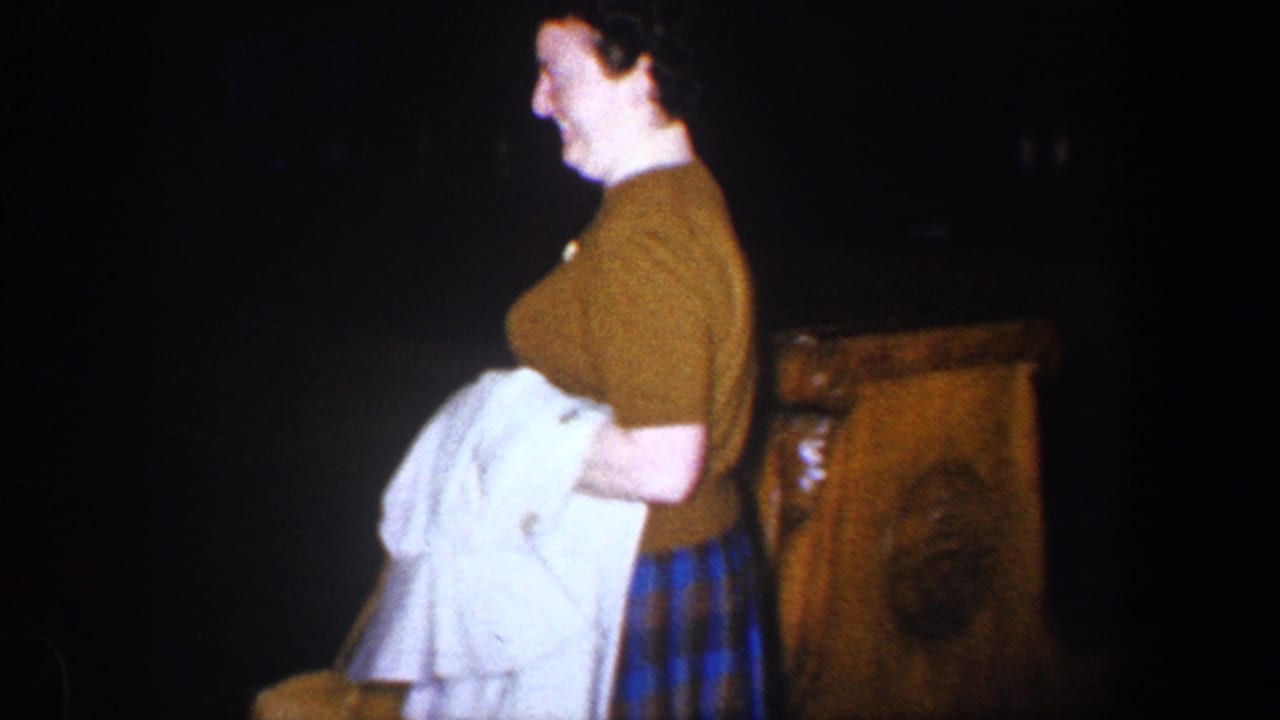 Woman in a brown sweater holds white clothing beside a leather armchair in a dark room.