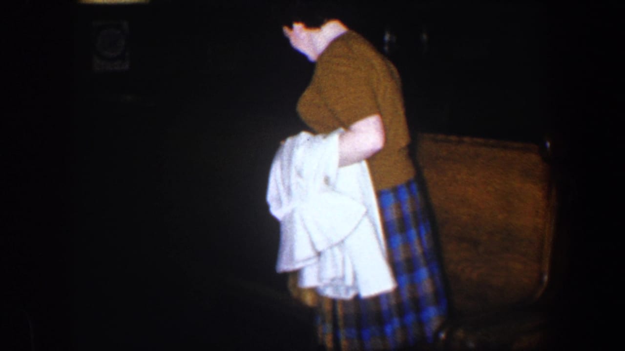 The woman turns slightly in the dim interior, holding a white garment beside a leather armchair in the 1960 Baltimore clip.