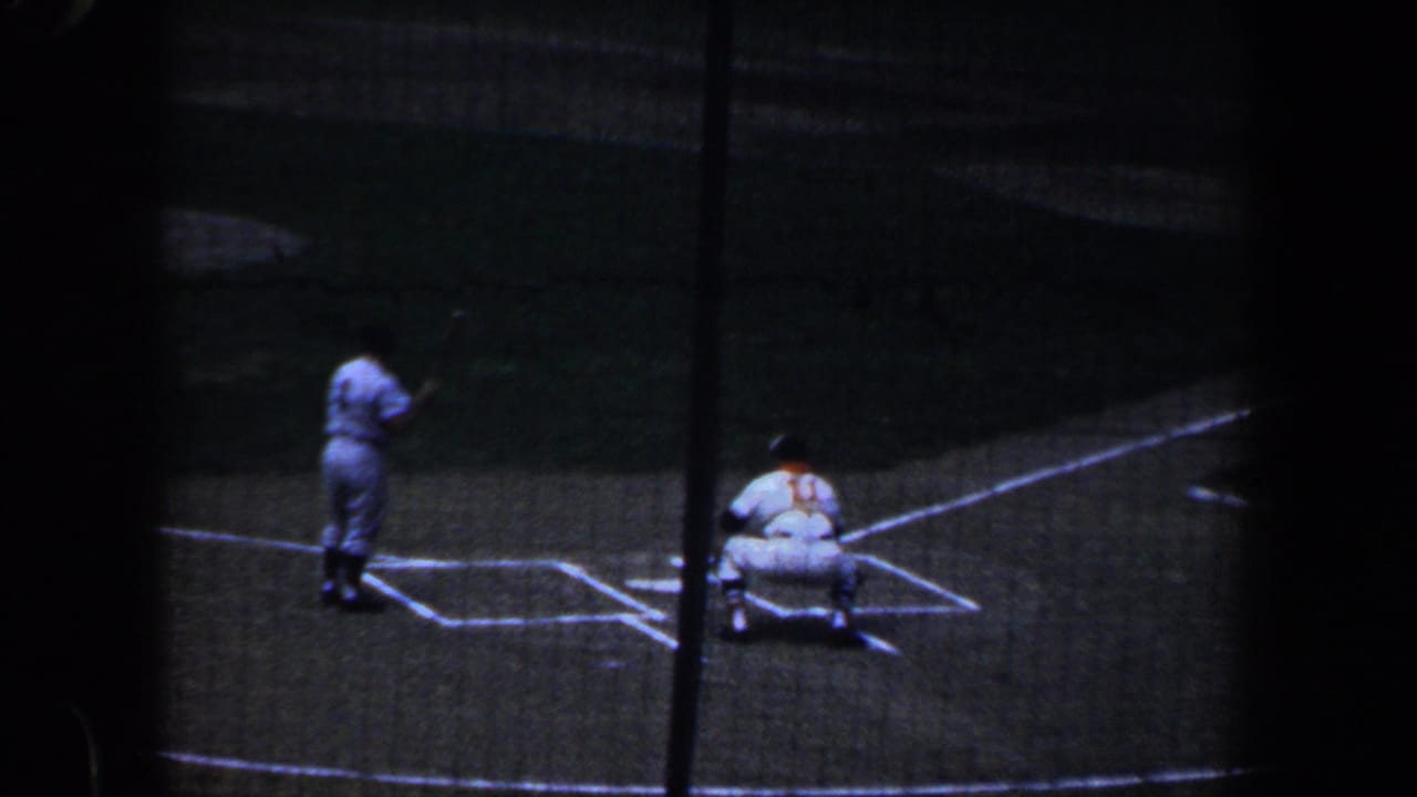 1960 Baltimore baseball game viewed from the stands through protective netting, with a batter near home plate and a catcher crouched behind.