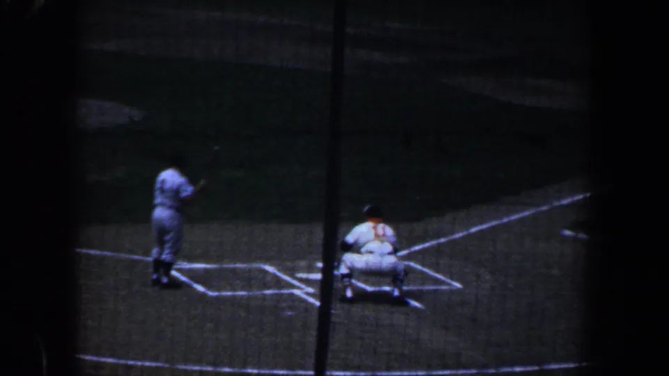 1960 Baltimore baseball game viewed from the stands through protective netting, with a batter near home plate and a catcher crouched behind.